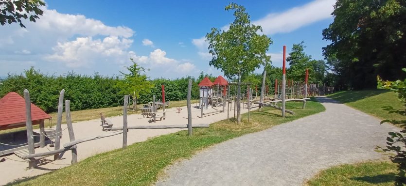 Spielplatz mit Geräten, Sandfläche und Bäumen unter blauem Himmel.