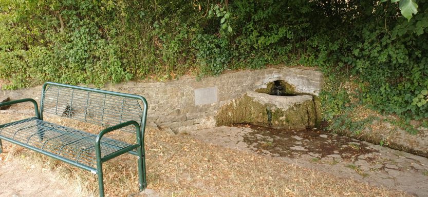 Historic Ittlinger Brunnen in Sinsheim-Weiler: Rustikaler Sandsteinbrunnen mit plätscherndem Wasser, Gedenktafel und grüner Bank.
