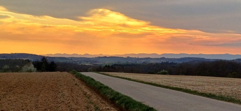 Ein asphaltierter Feldweg führt durch Ackerflächen unter einem golden leuchtenden Abendhimmel mit flachen Hügeln am Horizont.
