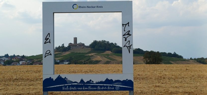 Großer Fotorahmen des Rhein-Neckar-Kreises auf einem Feld mit Blick auf die Burg Steinsberg in Sinsheim-Weiler.


